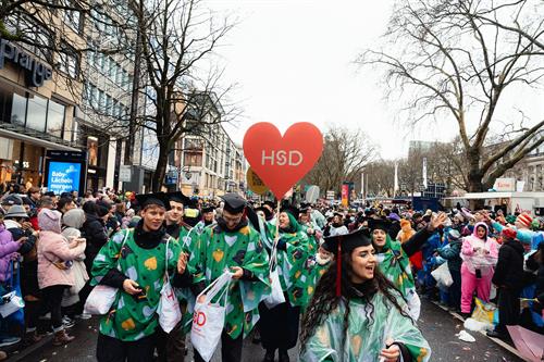 Studierende und Mitarbeitende der Hochschule Düsseldorf ziehen beim Rosenmontagszug durch die Düsseldorfer Innenstadt. In grünen Kostümen mit Doktorhüten tragen sie Wurfmaterial; über der Gruppe schwebt ein rotes Herz mit der Aufschrift „HSD“.